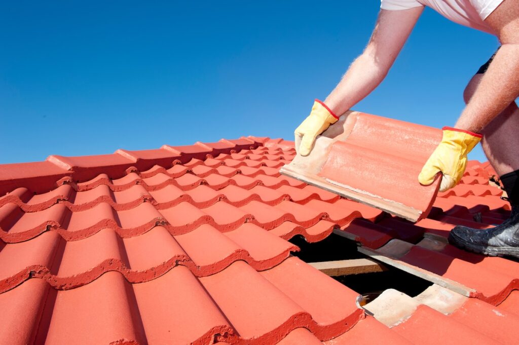 Roof repair, worker with yellow gloves replacing red tiles or shingles on house with blue sky as background