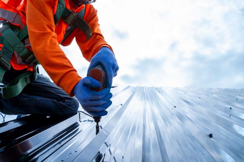 Close up photo of Professional and qualified roofer in protective uniform wear use electric drill to install the metal sheet on the new roof of new modern building construction.